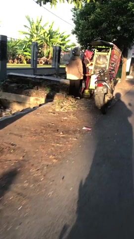 The media file shows a street scene in Ngampelsari, Indonesia, during what appears to be the late afternoon or early evening, given the long shadows cast on the ground and the warm, golden light. The weather seems clear and sunny.

The primary subjects are two individuals and a motorcycle. One person, wearing a helmet and a tan jacket over a red top and dark pants, is standing near the back of a motorcycle. This individual appears to be interacting with another person who is partially obscured and seems to be wearing a red outfit and a helmet. The motorcycle itself is a scooter, possibly a delivery vehicle, with a large, enclosed box mounted on the back. This box has graphics on it, but the text is not clearly legible. The motorcycle is parked on the side of a paved road.

The scene is set along a roadside with a shallow, debris-filled drainage ditch to the left of the road. Beyond the ditch, there is a low wall or fence, and behind that, lush green vegetation, including banana trees. The overall setting suggests a semi-urban or suburban environment in Indonesia, characterized by tropical foliage and a generally modest infrastructure.

The activity taking place appears to be a brief stop or interaction, possibly related to a delivery or a break. The posture of the individuals suggests they are engaged in conversation or completing a task. There are no strong indicators of emotion visible, but the scene is peaceful and ordinary. The lighting creates a pleasant, warm ambiance. Visible text on the motorcycle box is blurry and unreadable.
