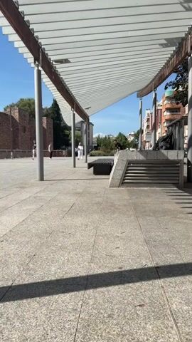 A young man wearing a black t-shirt and brown pants is skateboarding. He approaches a dark-colored park bench from the left, jumps onto the bench, and rides across it on his skateboard. He then jumps off the bench, and the skateboard rolls away. The man continues to walk forward, looking at the camera. The scene takes place outdoors on a paved area under a white slatted canopy. In the background, there are trees, buildings, and a few other people walking. The sky is clear and blue.