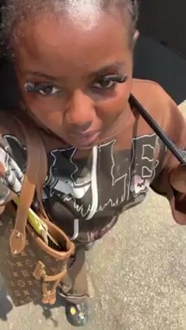 A young woman stands on a paved surface, possibly a sidewalk or street, holding a black umbrella slightly above her head to provide shade in bright sunlight. She is wearing a brown and black patterned zip-up sweatshirt over a brown shirt or pants. A brown Louis Vuitton purse hangs from her left shoulder. The woman is looking towards the camera and then turns her head to the right. She has braids in her hair and is wearing prominent false eyelashes. The surrounding environment appears to be outdoors on a sunny day.