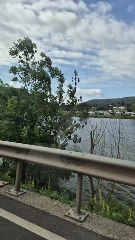 The video captures a scenic view from a moving vehicle, looking out across a body of water with hills and trees in the background. A metal guardrail runs along the side of the road in the foreground. The sky is partly cloudy. The water has ripples and reflects the sky. Distant white structures, possibly buildings or tents, are visible on the far bank of the water. The vehicle's side mirror is briefly visible on the left side of the frame as the camera pans. The overall impression is one of a peaceful, natural landscape.