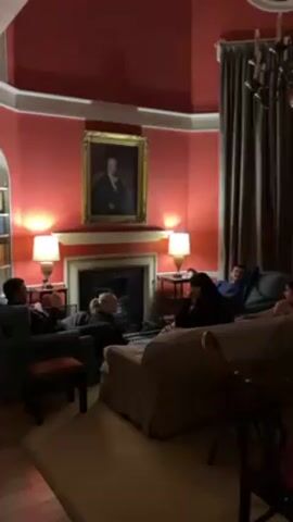 A group of five or six people are gathered in a cozy living room. The room is painted a deep red on the walls, with a white cornice. A portrait hangs above a fireplace. Two lamps on side tables flank the fireplace. A bookshelf is visible in an alcove to the left. The group is scattered among several armchairs and sofas. They appear to be engaged in conversation. A chandelier hangs from the ceiling.