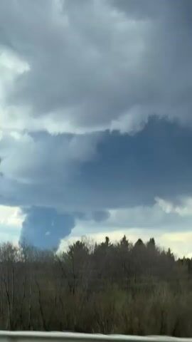 The video, filmed from a moving vehicle, captures a large, grey, plume-like formation rising into the sky above a dense forest. The sky is heavily overcast with dark grey clouds, but there's a lighter area visible towards the horizon. The plume itself is columnar, thick, and appears to be a significant event, possibly a large smoke cloud from a fire or an industrial emission.

In the foreground, a white road barrier is visible, indicating the viewer is on a road or highway. Beyond the barrier, a line of tall, bare-branched trees initially obscures part of the view, quickly blurring as the vehicle moves. Behind these, a solid green treeline, likely a coniferous forest, stretches across the midground. Power lines or utility poles are also briefly seen intersecting the view as the vehicle passes. The movement of the vehicle causes blurring in the foreground and midground, while the large plume in the distance remains relatively clear. The scene is set in a rural or forested area, with the location context provided as Lipaki, Russia. No people or visible text are present in the video.