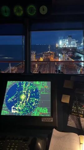 This video shows the interior of a ship's bridge at night. A large radar screen displays a circular image with many green and yellow blips, indicating radar contacts. To the right of the radar screen are various buttons and controls. Above the radar screen, across the top of the windows, are several circular gauges with illuminated needles. The windows look out onto the dark sea and the lights of other ships and a port facility. The overall impression is one of a working vessel on watch at night.