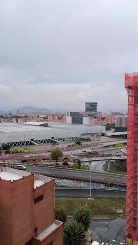 The video shows a city skyline under a cloudy sky. In the foreground, there is a red brick building and a telecommunications tower. In the middle ground, there is a large, modern convention center with a white roof and a complex road system with cars driving on it. In the background, there are many buildings and hills. The camera is panning slowly to the right.