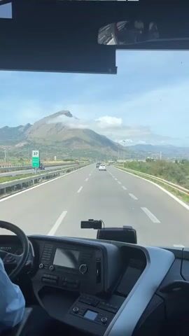 The video captures a first-person perspective from the driver's seat of a bus, looking forward through the windshield at a highway. The highway is straight and has multiple lanes, with white markings on the asphalt. Several cars are visible on the road, driving in both directions. In the distance, a large mountain with some clouds near its peak dominates the landscape under a clear blue sky. On the right side of the road, there's an exit with a white sign indicating "Imbarco". The bus dashboard is visible at the bottom of the frame, showing various controls and a steering wheel on the left, partially obscured by a person's arm.