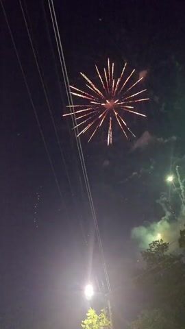 A fireworks display illuminates the night sky with bursts of light and color. A series of explosions in pink and white hues light up the darkness. Smoke plumes rise from the explosions. The scene is captured from a low angle, with power lines and a streetlight visible in the foreground. Trees are silhouetted against the vibrant sky.