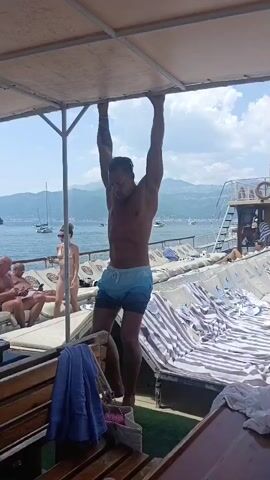 A shirtless man with muscular abs and arms performs hanging leg raises. He is holding onto a horizontal bar under a canopy. He is wearing blue and white swim trunks. In the background, there is a body of water with boats and a shoreline with mountains. There are lounge chairs with striped towels and people relaxing on them.
