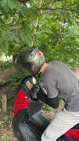 A person wearing a helmet and a grey shirt is seen on a red and black scooter. The person appears to be checking something on the scooter’s console. The scene is outdoors, surrounded by lush green foliage and trees. There's a fallen log on the left side of the frame. The lighting suggests it is daytime.