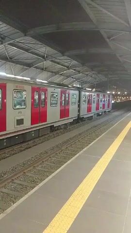 The video shows a subway train moving away from the perspective of someone standing on a platform. The train is silver with red doors and a few windows. The platform is gray with a yellow safety line. The station has a metal roof structure. The train is moving quickly away from the camera.