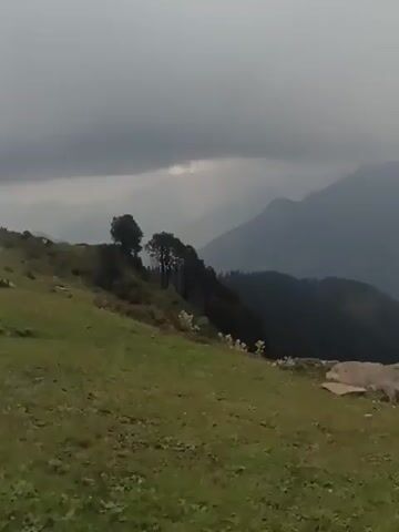 The video shows a panoramic view of a mountainous landscape. In the foreground, there is a grassy meadow with scattered rocks and a few small yellow flowers. A few trees are visible on the left side of the frame. In the middle ground, there are rolling hills covered in greenery. The background consists of layers of dark, misty mountains fading into the distance. The sky is overcast with dark, dramatic clouds, suggesting cloudy or possibly rainy weather. Towards the end of the clip, a few people can be seen sitting on the grassy slope in the middle ground, appearing to be enjoying the view. The camera slowly pans across the landscape.