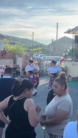 In an outdoor setting, likely a village street, a group of people are gathered. The focal point is a performance by two couples in traditional attire. One couple on the left consists of a man in a yellow shirt with a brown vest and dark pants, and a woman in a white top and a patterned skirt. The other couple on the right mirrors this setup. They are dancing in unison with energetic movements, their arms often extended. Spectators are positioned in the foreground and background, some seated and others standing. In the foreground, three women are facing the camera, two of whom appear to be conversing. Behind the dancers, cars are parked along the street, and houses with tiled roofs line the background. The sky is visible with clouds, and a flagpole with a flag, possibly national, is present. There is a sense of community celebration.