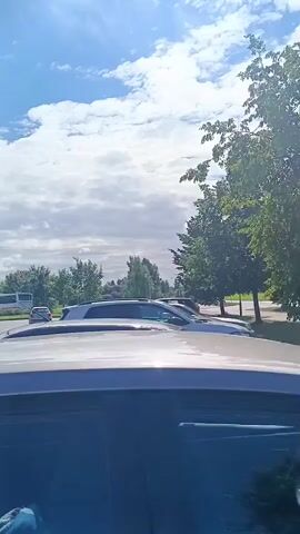 A daylight exterior shot shows a parking lot with several cars. In the foreground, the roof of a gray car is visible, and the interior of the car is partially visible, with a person wearing a blue shirt and jeans in the driver's seat. In the background, a white van is parked, and a white bus is visible on a road to the left. Beyond the parking lot, there are trees and a building. The sky is blue with white clouds.