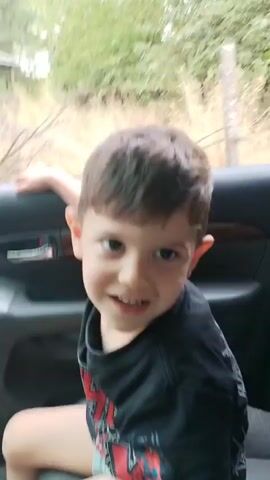A young boy is sitting in the back seat of a car. He is wearing a black t-shirt and blue shorts. He is smiling and looking at the camera. The car is moving. The background is blurry, suggesting movement. The boy's left arm is resting on the car door.