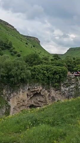 The video shows a scenic view of a gorge with lush green hills on either side. In the foreground, there is a grassy slope with some yellow flowers. The gorge itself is deep, with rocky cliffs and dense vegetation. On the far side of the gorge, there is a bridge or platform with people standing on it, some of whom are waving Russian flags. Further back, on the hillside, a large yellow construction vehicle, possibly a bulldozer, is visible amidst the trees. The sky is overcast with dramatic clouds. The camera pans slightly to the right throughout the short clip, revealing more of the landscape and the distant construction equipment.