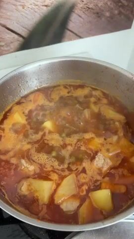 A close-up shot shows a large metal pot filled with a simmering stew. The stew contains chunks of potatoes, carrots, peas, and meat in a reddish-brown broth. A wooden spoon is visible, stirring the contents of the pot, causing some of the stew to splash around the edges. The pot sits on a stovetop, and steam rises from the dish. In the background, a wooden-patterned surface is visible, possibly a backsplash or wall. The lighting suggests an indoor setting, likely a kitchen.