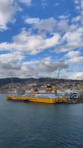 The scene is a port with several large yellow and white ferries docked. The ferries are labeled "Corsa Sardinia Ferries." Two tall cranes are visible near the ships. The background includes a densely populated city sprawling up a hillside, with mountains in the distance. The sky is blue with scattered white clouds. The water is dark blue, reflecting the sky.