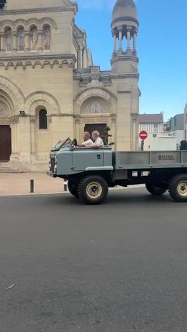 The video shows a grey, vintage flatbed truck with two people in the driver's seat. The truck is driving on a paved street in front of a large, ornate building that appears to be a church or cathedral, with arched doorways and decorative facades. The building has a dome-shaped tower on its right side. To the left of the church, there are multi-story buildings with windows and balconies. Trees line the sidewalk on the left side of the street. The sky is blue with some clouds. The truck has large, off-road style tires and a flatbed in the back carrying a white object. The truck has "FILTRES SOPHIA" written on the side of the bed. The truck is moving away from the camera. The video is shot from a slightly low angle, capturing the street and sidewalk in the foreground.