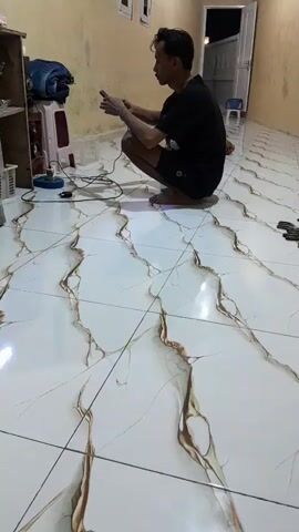 A video shot from a low angle shows a man in a black t-shirt and shorts kneeling on a tiled floor. He is holding a mobile phone in his hands and appears to be engaged with it. The room has yellow walls and a white tiled floor with a marble-like pattern. To the left, there is a wooden shelf unit with various items, including what looks like a fish tank, plastic crates, and other miscellaneous objects. A red and white stool is also visible near the shelf. In the background, there are doorways and windows, suggesting an indoor setting, possibly a home or a shop. The lighting is dim, indicating it could be evening or nighttime. Another person's legs and feet are visible at the far left of the frame, indicating the presence of at least two people. The scene appears to be casual and ordinary, with no obvious signs of distress or celebration. There is some text visible on a white crate on the floor, appearing as "SH". The location context is Panjalin Kidul, Indonesia.