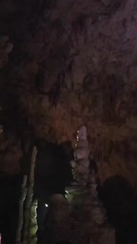 This is a short video showing a cave formation. The video begins with a close-up of a stalagmite in a dimly lit cave, with other formations visible in the background. The lighting then shifts, becoming darker, and the video ends with what appears to be a view of the cave floor or possibly water, with some faint reflections. No people or specific actions are clearly depicted.