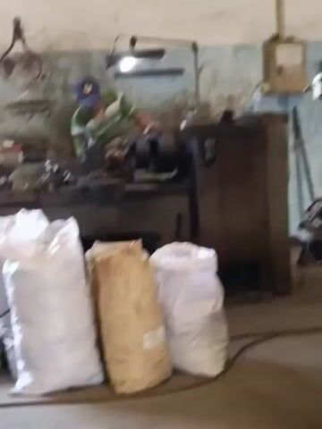 A man wearing a green and white striped shirt and a blue baseball cap is operating a metal lathe in a workshop. He is standing and appears to be machining a metal object. In the foreground, there are several large sacks, some white and one appearing to be filled with grain or straw. The workshop environment includes various tools and equipment mounted on the walls and benches, with a workbench and the lathe machine dominating the central area. The lighting suggests an indoor setting, possibly a garage or industrial space.