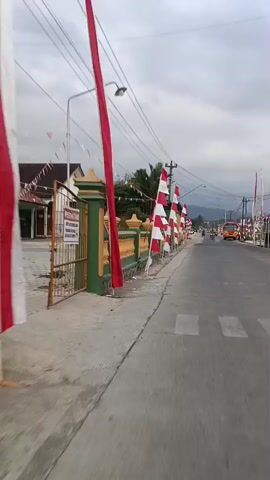 The video shows a street lined with red and white Indonesian flags. The flags are attached to poles and are placed along the sidewalk and a low wall. A green and yellow fence is visible on the left side of the road. A paved road stretches into the distance, with vehicles and people sparsely present. The sky is overcast. The perspective suggests the video is being filmed from a moving vehicle or bicycle.