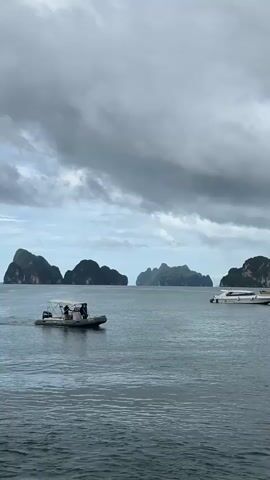 The video shows a wide shot of a body of water, possibly a bay or ocean, with several karst islands in the distance. The sky is overcast with dark clouds. In the foreground, a rigid inflatable boat (RIB) with a canopy is moving from left to right across the frame. Three people are visible in the RIB. To the right of the RIB, a white speed boat is stationary. The water is choppy, and there is a slight ripple effect on the surface. The overall mood of the video is somewhat dramatic due to the cloudy sky and the vast expanse of water.