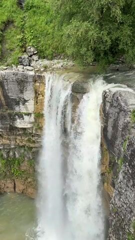 A video shows a wide, powerful waterfall cascading over a rocky cliff face. The water is white and foamy as it plunges into a greenish pool below. The surrounding cliffs are layered and appear to be made of stone with some vegetation growing on them. Lush green trees and foliage are visible at the top of the cliff and on the surrounding slopes. The perspective is from above and slightly to the side of the waterfall, looking down. The weather appears to be overcast or the lighting is diffused. The water flow seems continuous and substantial.