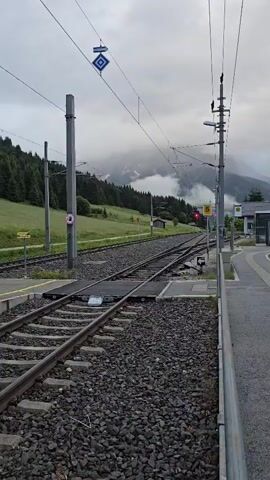 The video shows a train track intersection in a mountainous region. The tracks are surrounded by gravel and a grassy slope dotted with trees. In the background, misty mountains rise above the trees. Overhead, there are electrical wires for a train and utility poles. A red signal light is visible, indicating a stop. There are also various signs and a small building. The camera pans slightly, revealing more of the track layout and the surrounding landscape.