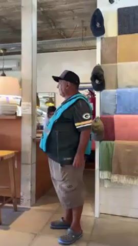The video shows a man standing in what appears to be a shop or store. He is wearing a Green Bay Packers shirt, khaki shorts, and grey Crocs. Over his shirt, he has on a bright blue vest. He also sports a two-toned black and grey baseball cap. The man is looking slightly to his right, seemingly engaged in a conversation. In the background, a woman is visible behind a counter.  The store has various blankets displayed on a shelving unit and a few hanging hats. The general atmosphere suggests a retail setting.