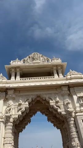 The video pans down through an ornate white archway, revealing a view of a large white temple with multiple spires in the distance. The temple has a fountain in front of it, and a few people can be seen near the fountain. The sky is blue with some clouds. The archway is highly decorated with intricate carvings and pillars. The ground in the foreground is paved with red and brown bricks.