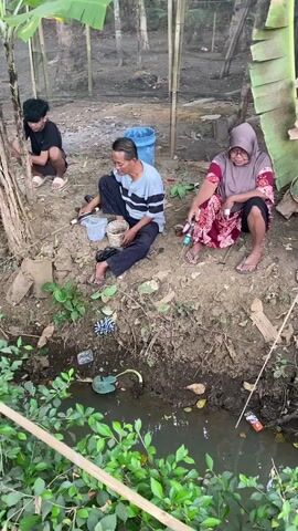 A group of people are gathered by a narrow, murky body of water. Several individuals are sitting on the muddy bank, while others are standing. One person, a woman wearing a headscarf and a patterned dress, is using a long stick to poke at something in the water. Another person, a man in a striped shirt, is sitting with a bucket next to him. Two children are sitting together, and a man wearing a red shirt is also present. The environment appears to be outdoors, with some vegetation and wooden structures visible in the background. The water appears to have some debris floating in it.