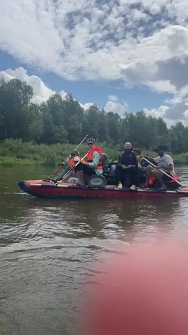 The video shows a group of people on a raft on a river. The raft is red and blue and has an outboard motor. The people are wearing life jackets and are holding paddles. The raft is moving through the water and the trees are visible on the shore. The sky is blue with some clouds.