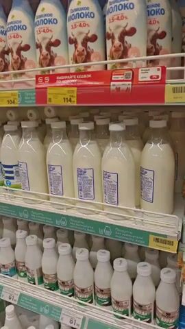 The video pans across shelves in a supermarket dairy aisle. Multiple brands of milk in various bottle and carton sizes are visible, predominantly white with blue, red, and yellow accents on the labels. Some bottles have blue and white striped labels, while others have white labels with red lettering. The shelves are well-stocked, and price tags are visible beneath some of the products. The overall environment appears to be a brightly lit retail space. There are no people visible in the frame.