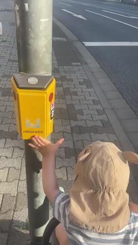 The video begins with a close-up of a yellow pedestrian crossing button on a metal pole, featuring a graphic of people crossing and the word "DRÜCKEN" (German for "push"). A child wearing a tan sun hat with ear-like flaps and a striped shirt is reaching towards the button with their right hand. The camera then pans to show a street scene in Nidda, Germany.

The environment appears to be a town or city with modern buildings, including a multi-story building with a pink facade and horizontal blinds, and another building with a traditional half-timbered style above shops. The street has crosswalk markings and traffic lights. The sun is shining brightly, suggesting it is daytime, and the light is somewhat hazy, possibly indicating early morning or late afternoon. The overall mood seems peaceful and ordinary, capturing a moment of a child preparing to cross the street. Visible text includes "DRÜCKEN," "HOLLYWOOD," and "nara." The presence of the pedestrian crossing button and crosswalks indicates an urban or suburban setting where pedestrian safety is a consideration.