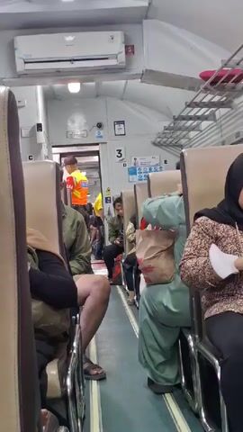 The video shows the interior of a train carriage. Passengers are seated on both sides of an aisle. Some passengers are wearing masks. At the end of the aisle, a person in a yellow and orange vest is visible near a doorway. An air conditioning unit is mounted on the ceiling. There are signs with numbers and text visible on the walls. The lighting appears to be artificial.