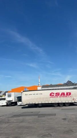 A freight truck with "CSAD LOGISTIK OSTRAVA" printed on its side is parked in an industrial area under a clear blue sky. A tall, striped smokestack is visible in the background, along with orange and white buildings. Another white truck is parked to the left of the freight truck.