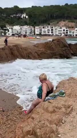 Here is a description of the video:

The video shows a woman sitting on a rocky outcrop at the edge of a beach. She is wearing a swimsuit and appears to be relaxing as waves crash around the rocks. In the background, there is a sandy beach with people and white buildings. There is a forest of trees behind the buildings. The weather looks overcast and slightly windy.