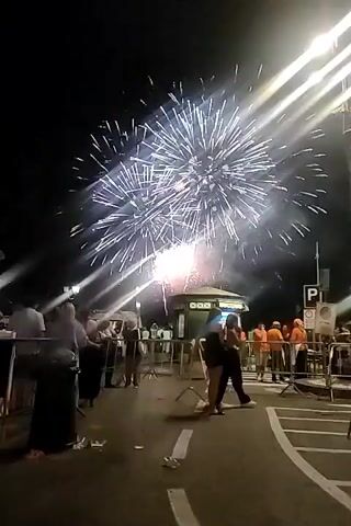 The video captures a night scene with fireworks exploding in the sky. The fireworks are white and appear to be bursting in multiple layers. In the foreground, a group of people are gathered, some standing behind a metal barrier, watching the display. Some people are silhouetted against the light of the fireworks. There is a small booth or structure in the middle ground, illuminated by the fireworks' light. The ground appears to be paved, with some white markings visible. The overall atmosphere suggests a celebratory event.