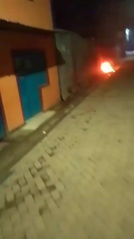 The video shows a narrow, brick-paved street at night. A small fire burns on the side of the road at the start of the video. The camera moves slowly down the street, revealing buildings on either side, a street light, and graffiti on one building that reads "MOTTO KNOWLEDGE IS POWER." The street is empty of people.