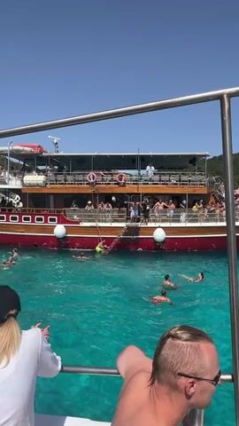 A boat with many passengers is anchored in clear turquoise water. People are swimming and enjoying themselves in the water. Some people are on the deck of the boat, while others are in the water. The boat has a red hull and a wooden deck. The sky is clear and blue. There are trees on the shore in the background.