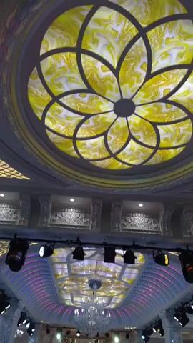 The video shows a lavishly decorated hall, likely for an event like a wedding. The hall features a grand ceiling with ornate patterns and colored lighting, a central chandelier, and multiple spotlights. A long, decorated aisle runs down the center of the room, flanked by abundant floral arrangements. People are present, some standing by the sides, while others are in the distance, possibly setting up or preparing for the event. One individual in the center of the aisle appears to be operating a camera or similar equipment. The overall impression is one of opulence and anticipation.