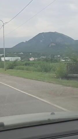 This video shows a view from a vehicle moving along a road. On the right side of the road, there is a ditch with tall green grass and some bushes. Beyond the grass, there are scattered houses and buildings, and a large mountain in the background. The mountain is covered in trees, with some lighter patches of exposed rock or soil on its slopes. Several utility poles with wires are visible along the road. The sky is overcast. The camera is angled slightly downwards, showing the dashboard or hood of the vehicle in the foreground. The movement of the vehicle causes a slight blur in the video.