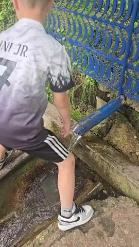 In this short video, a young boy is playing in a natural spring. He is wearing a grey and black t-shirt with a white number on the back, black shorts with white stripes, and grey and white sneakers. He is crouching near a blue pipe that is dispensing water, and he is splashing the water with his hands. The boy looks happy and excited as he plays in the water.

The scene appears to be outdoors in a natural setting with trees and greenery in the background. There is a blue metal fence with a decorative pattern behind the boy, and a wooden structure in front of him. The weather appears to be pleasant, as the boy is comfortably playing in the water.

In the background, there is a sign with some text, but it is not legible enough to read. The overall mood of the video is joyful and playful, capturing a moment of childhood fun in nature.