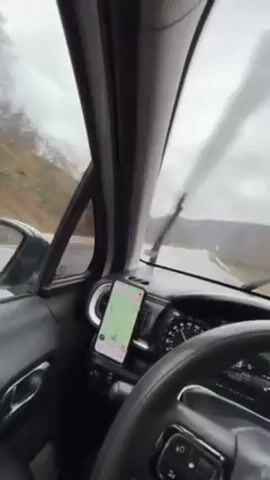 From the driver's seat, a vehicle navigates a rain-slicked road under a cloudy sky. The dashboard shows a smartphone mounted, displaying a map application. Wipers intermittently clear the windshield as the car progresses. The view ahead reveals a straight stretch of road bordered by trees and distant hills veiled in mist. The speedometer indicates 73, and the driver's hands are visible on the steering wheel.