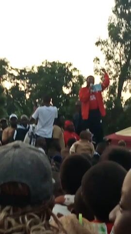 The video shows a group of people gathered outdoors, possibly for a religious service or a community event. A person in a red jacket is standing and speaking, holding a book or pamphlet. The crowd is seated or kneeling, facing the speaker. The background features trees and a tent or canopy. The lighting suggests it is either early morning or late afternoon. The people in the crowd appear to be diverse in age.