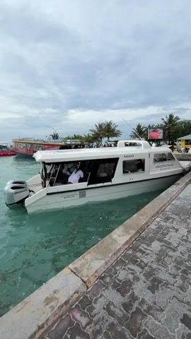 The video shows a white boat named "PASAKA" of the "Maldives Fellas" company docked at a concrete pier. There are people visible inside the boat. A Honda motor is attached to the back. The water is a light turquoise color with seaweed visible. In the background, there are other boats, palm trees, and a billboard. The sky is overcast with gray clouds.
