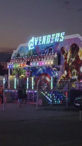 The video shows an Avengers-themed amusement park ride at dusk. The ride is brightly lit with colorful lights that flash and change. The artwork on the ride features characters from the Avengers movies, including the Hulk, Captain America, Black Widow, and Iron Man. People are seen standing around the ride and on the platform of the ride. The sky in the background is a deep purple, indicating that the sun has set. The overall atmosphere is festive and exciting.
