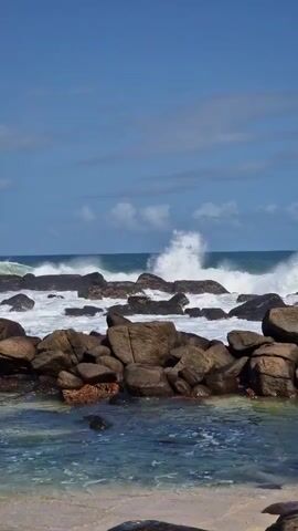 The video depicts a coastal scene on a bright, sunny day. Large, dark-colored rocks are prominently situated in the foreground and midground, forming a barrier against the ocean.  Waves crash against these rocks, creating white foamy splashes. Beyond the rocks, the ocean stretches to the horizon under a clear, blue sky with scattered white clouds. The water in the foreground is shallow and relatively clear. A sandy beach is visible at the bottom of the frame. There are no people or animals visible in the video. The camera is mostly static, providing a steady view of the ocean and rocky coastline.