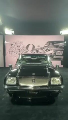 A black Toyota Century is on display. The car is facing the camera with its headlights and grill prominently visible. Behind the car, there is a pink wall with an illustration of the car's engine. The car is sitting on a dark, carpeted platform.  The lighting is bright, highlighting the car's features.
