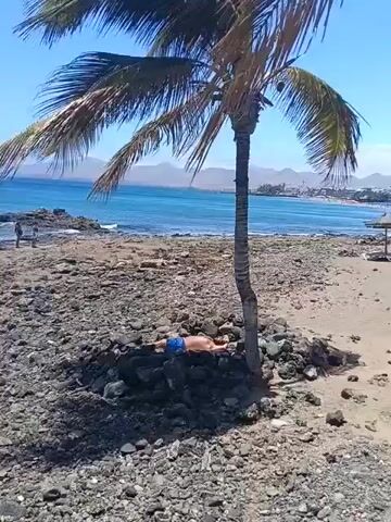 A man in blue shorts is seen lying on a bed of dark rocks under the shade of a palm tree on a sunny beach. He appears to be resting or sleeping, with a water bottle visible near his head. The foreground and middle ground of the scene are dominated by numerous grey and black volcanic rocks, characteristic of the area. In the background, the clear blue ocean with gentle waves laps against the shore. Distant land formations or mountains are visible across the water. Further along the beach, two other people are walking near the water's edge. The sky is bright and clear, indicating a sunny day. This rocky coastal scene is located in Puerto Del Carmen, Spain.
